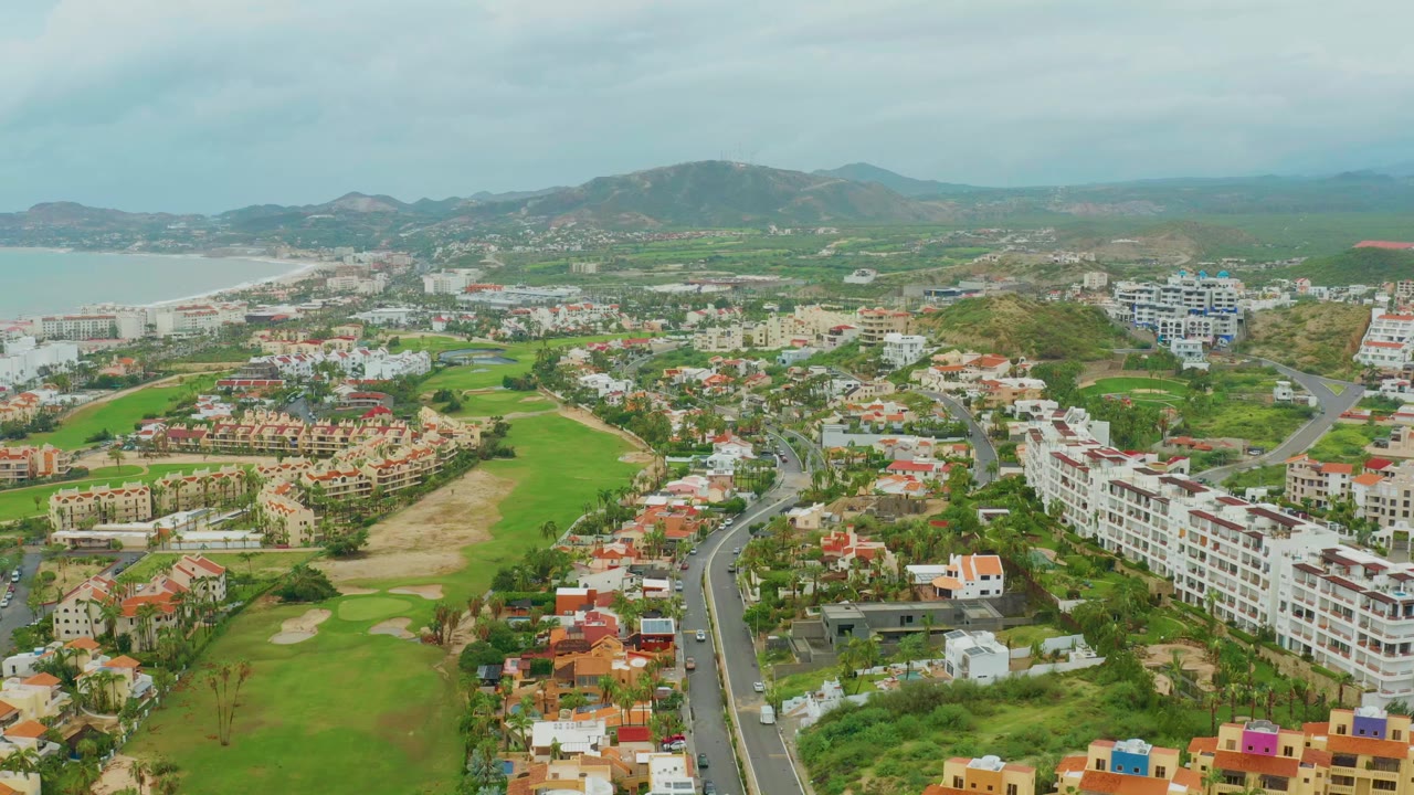 Aerial frame in San Jose del Cabo, Baja California Sur showing the road running up from foreground homes toward white buildings, golf fairways, distant coastline, hills, and no visible people.