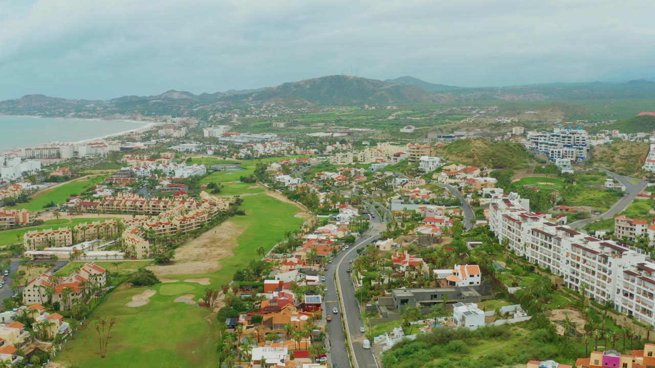 Wide drone view of coastal residential streets, green course land, and mountains.