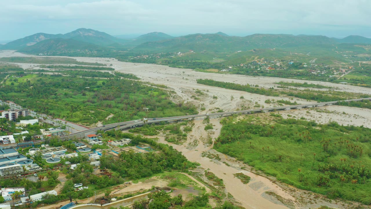 A wide aerial frame in Baja California Sur, Mexico shows a muddy flooded river spreading across a green valley, a road bridge in the midground, distant mountains, and no visible people.