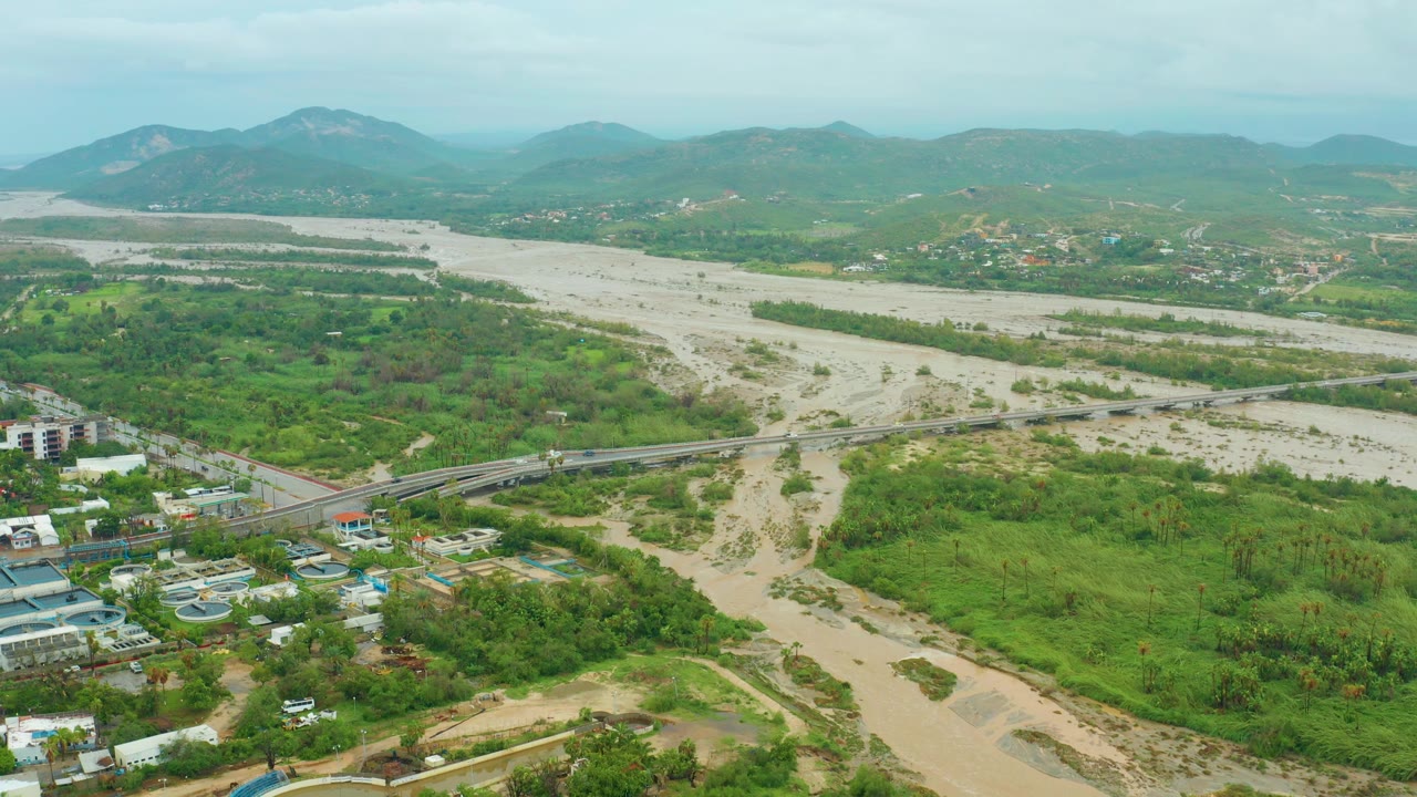 The second frame keeps the aerial view over Baja California Sur, Mexico, with the muddy river channel, bridge, green vegetation, small buildings near the lower edge, and no visible people.