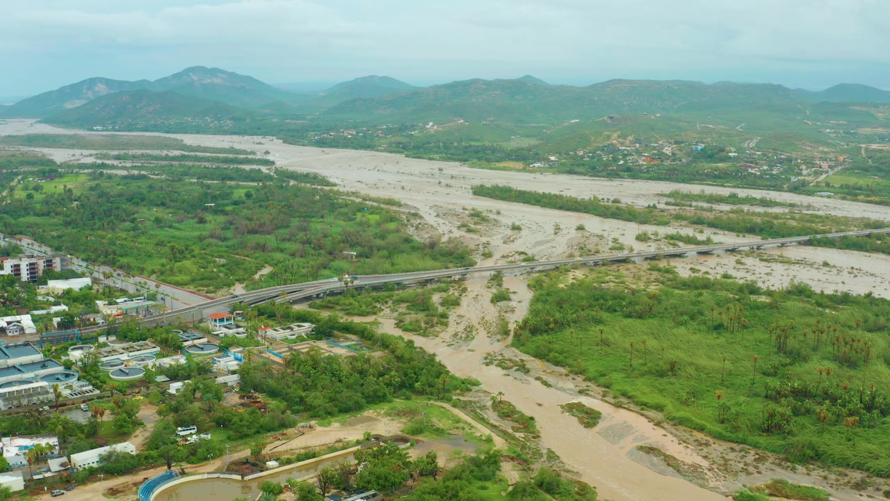 Flooded river valley with bridge and green hills under overcast sky.