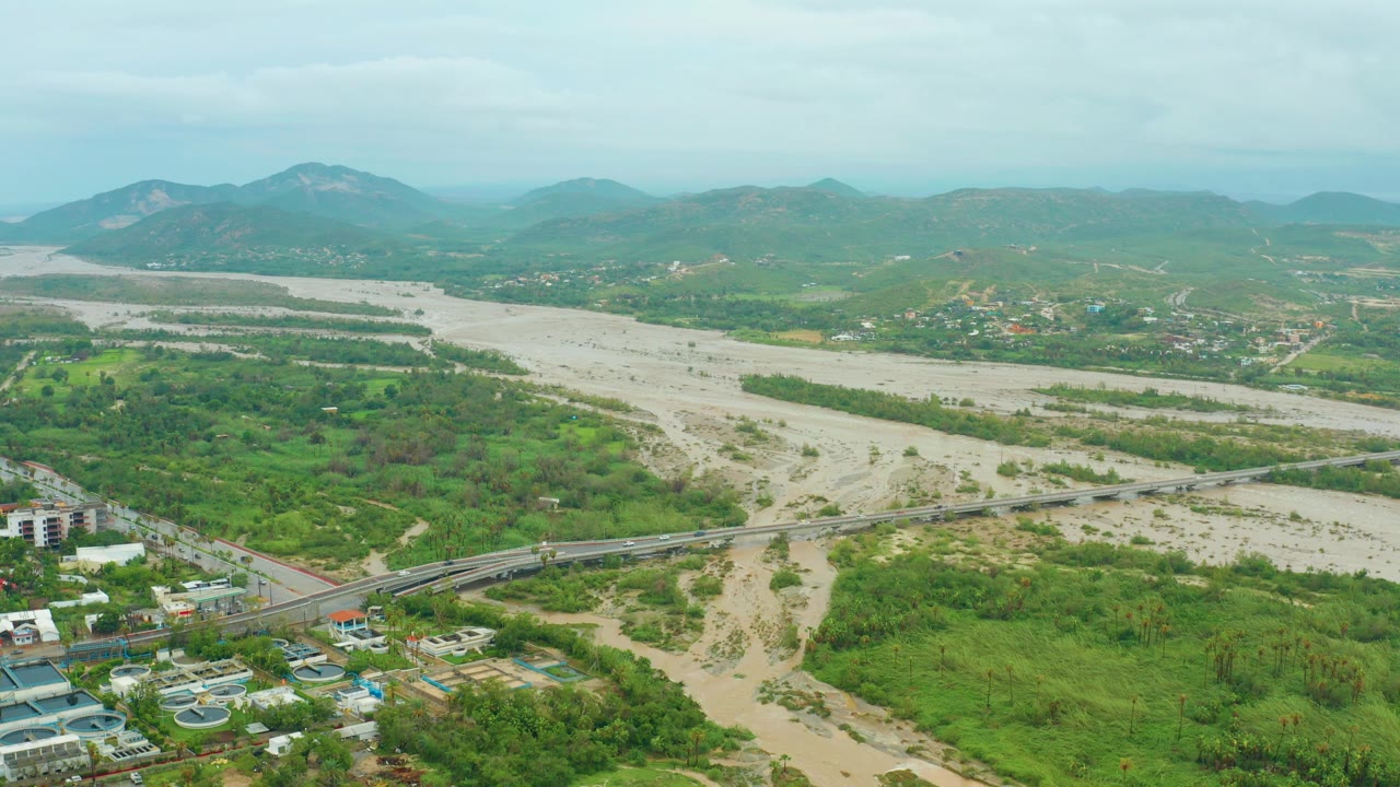 At 0:00, a wide aerial view in San Jose del Cabo, Baja California Sur, Mexico shows a muddy river valley, a bridge, green vegetation, small buildings, distant mountains, and no visible people.