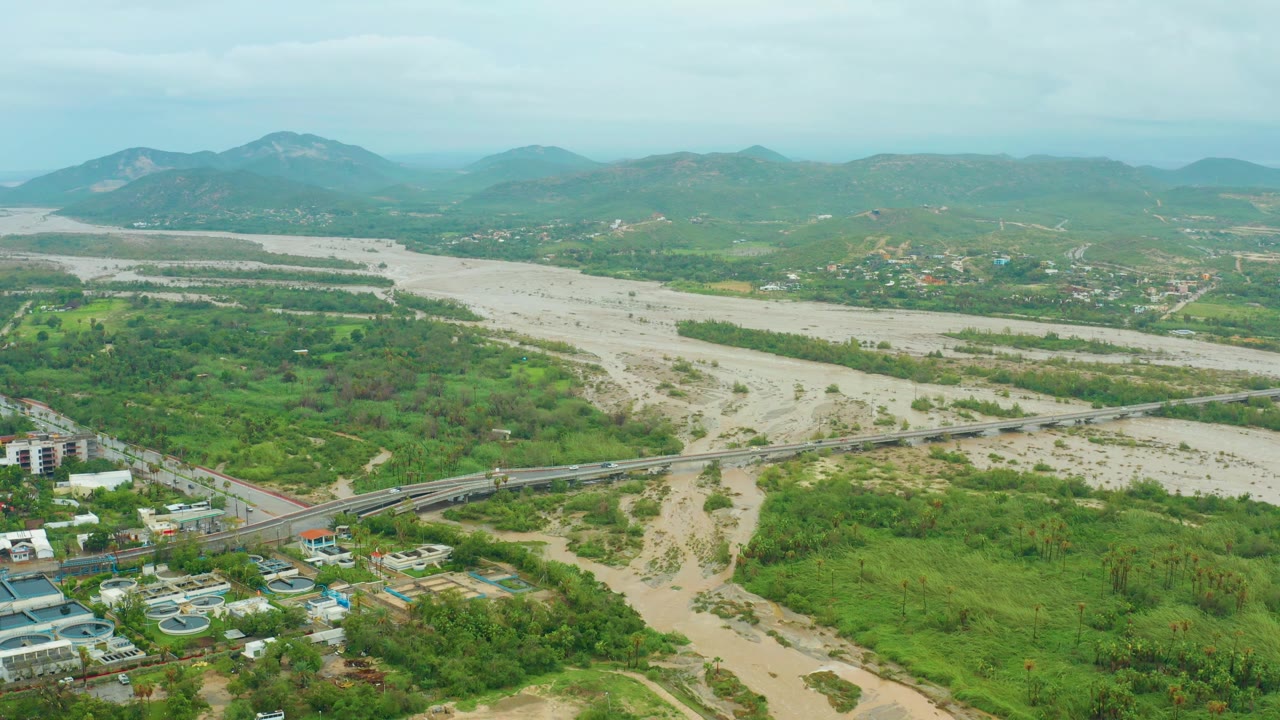 Wide aerial shot of a receding muddy river, green vegetation, bridge, nearby buildings, and low mountains.