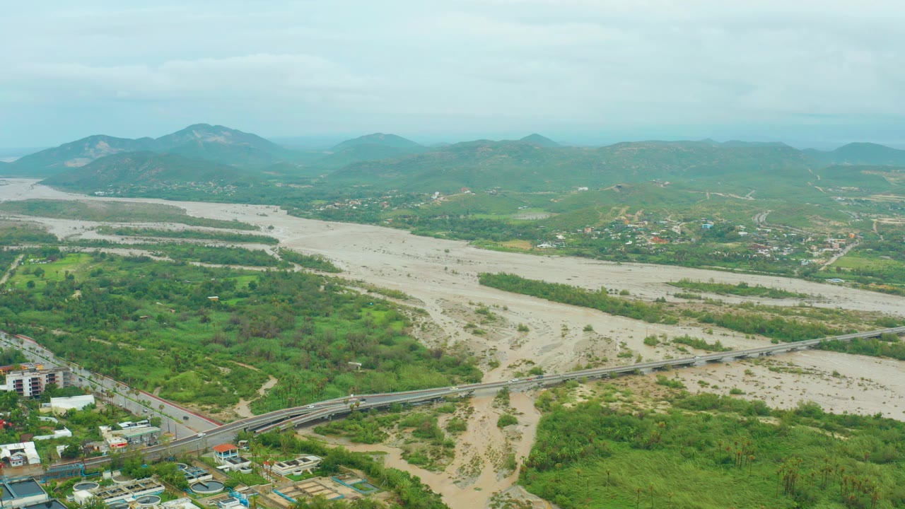 Opening aerial frame over San Jose del Cabo BCS, Mexico, showing a wide muddy river, green valley vegetation, a road bridge with small vehicles, distant buildings, mountains, and no visible people.