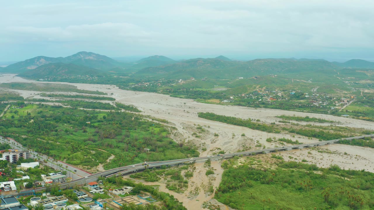 Aerial frame from San Jose del Cabo BCS, Mexico, showing the tan floodwater channel, green banks, bridge and road activity in the foreground, distant hillside buildings, mountains, and no visible people.