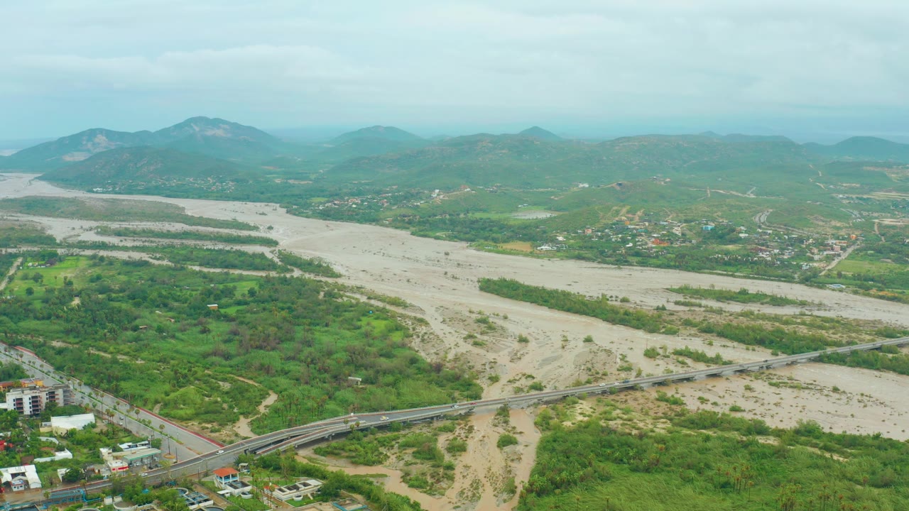 Wide aerial landscape with flooded river channel, road bridge, hills, and buildings.