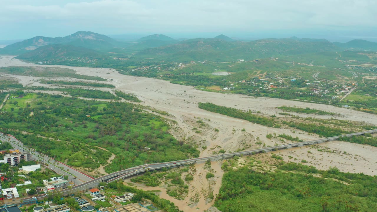 Aerial frame of the San Jose del Cabo valley with the sediment-colored riverbed spanning the middle, green banks, a road bridge near the lower edge, small buildings nearby, distant mountains, and no visible people.