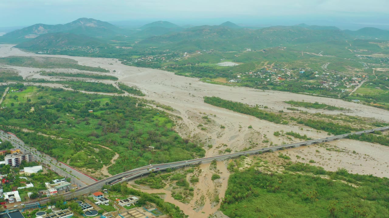 Opening aerial frame over San Jose del Cabo, Baja California Sur, with a muddy riverbed cutting through green vegetation, a bridge in the foreground, hazy mountains beyond, and no visible people.