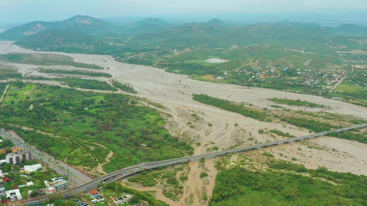 Aerial view in San Jose Del Cabo showing a muddy river channel, a road bridge near the lower frame, green vegetation, scattered buildings, hazy hills, and no visible people.