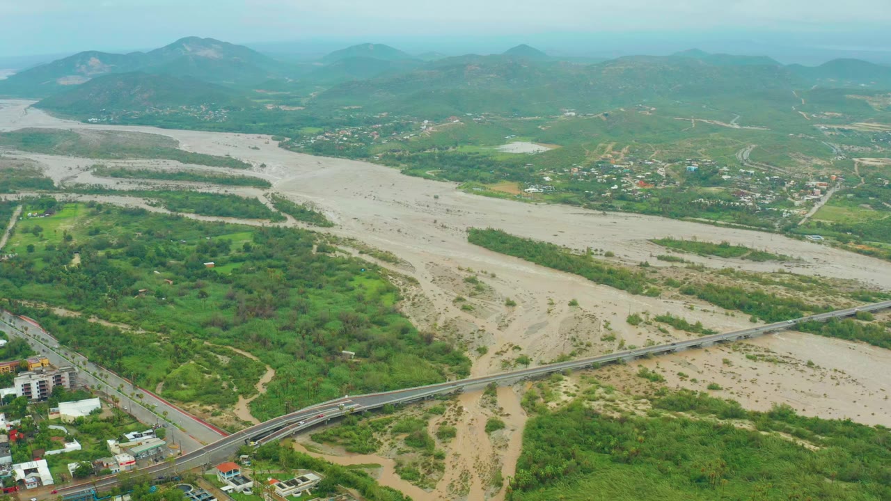 Wide drone frame over San Jose Del Cabo with pale muddy water spreading through the riverbed, a bridge crossing the lower midground, green banks, small buildings, distant mountains, and no visible people.