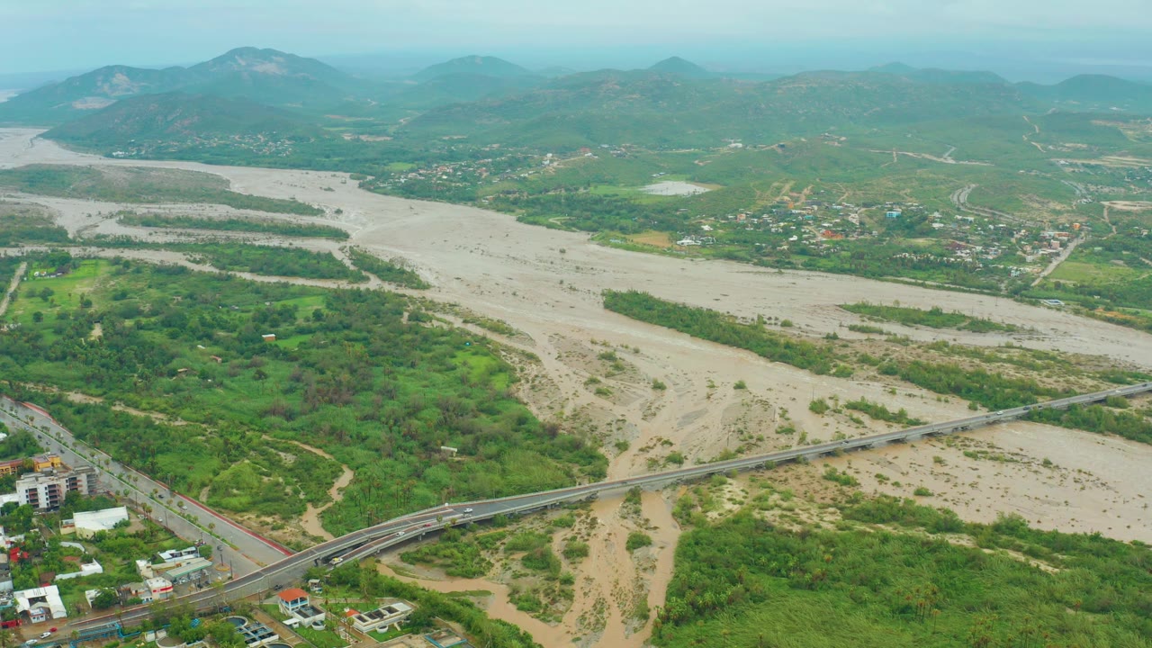 Aerial landscape with muddy riverbed, road bridge, greenery, and mountain backdrop.