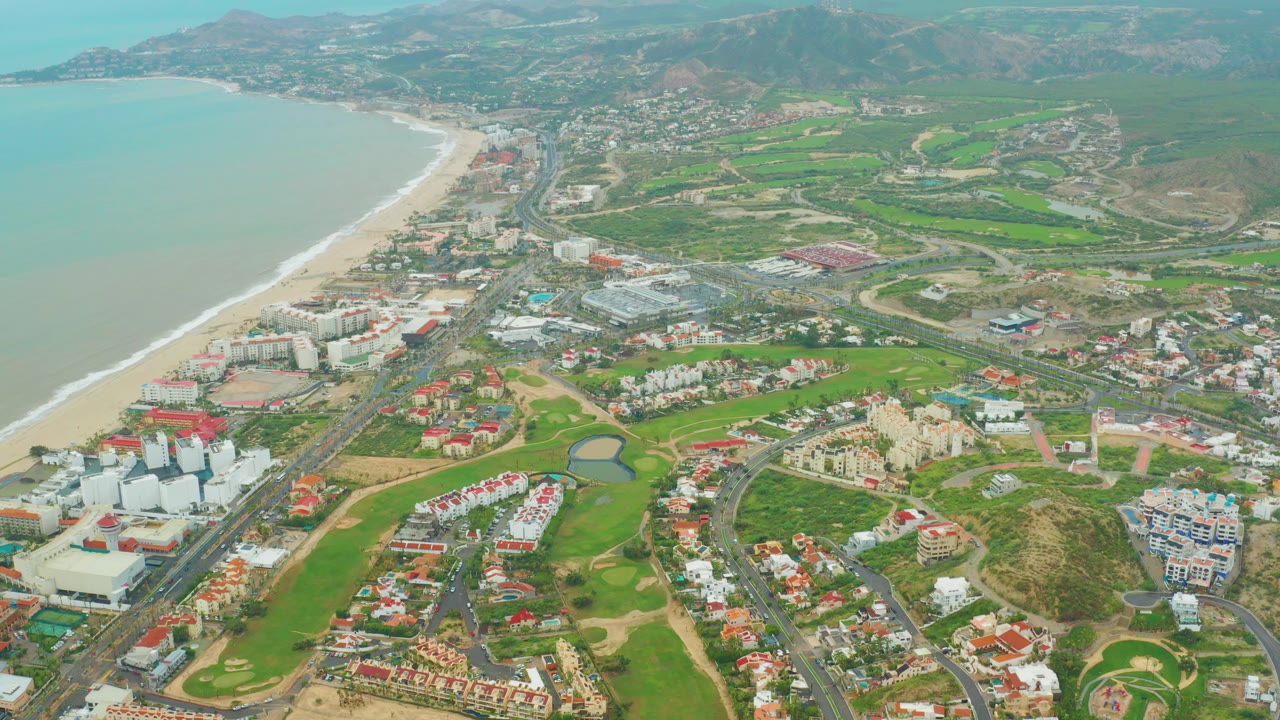 Aerial view over San Jose del Cabo with pale ocean and sandy beach at left, golf fairways and resort buildings below, hills in the distance, and no visible people.