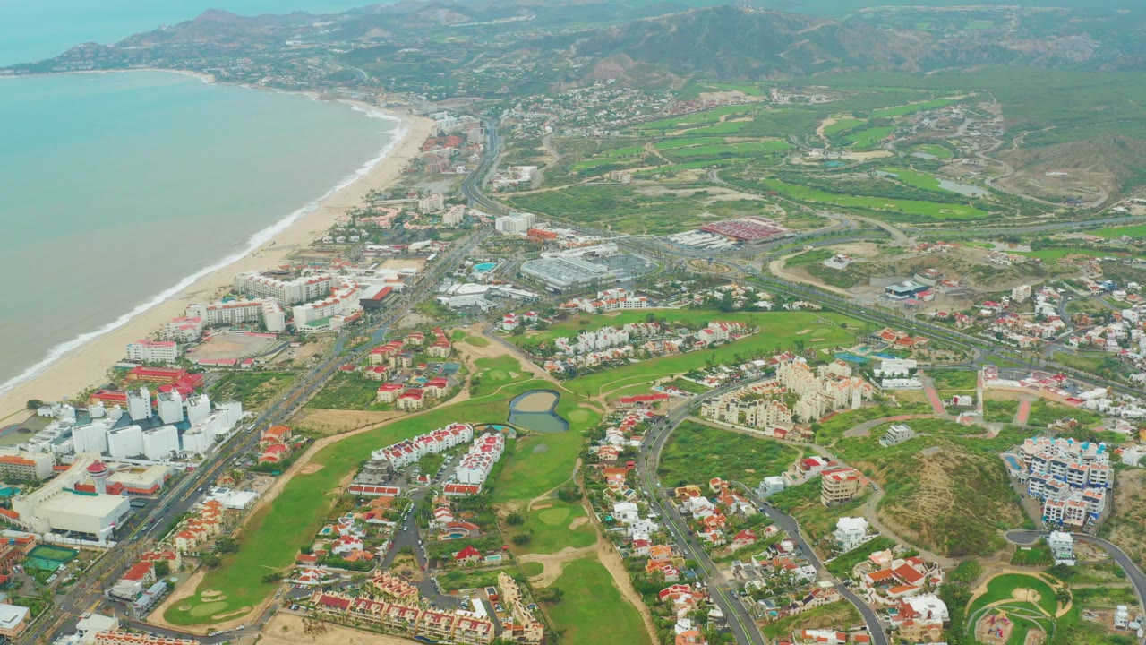 Wide aerial over San Jose del Cabo showing the beach curving along the left edge, green resort grounds and red-roof residences in the center, mountains beyond, and no visible people.