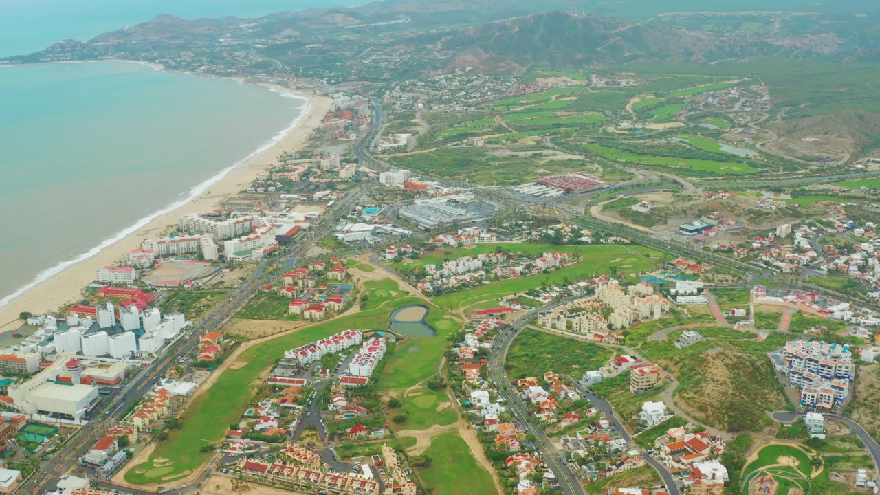 Drone-height view of San Jose del Cabo coastline with ocean surf on the left, resort blocks and red-roof homes below, golf greens through the center, distant hills, and no visible people.