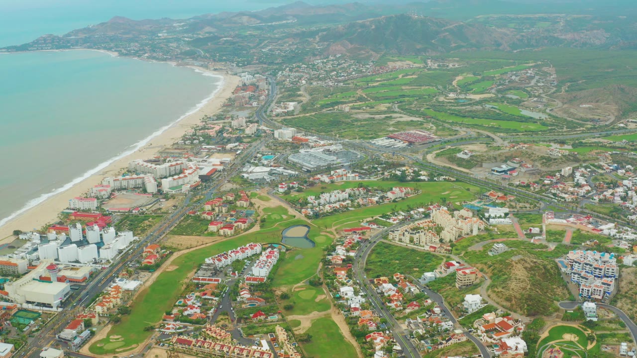 Coastal aerial with ocean, beach hotels, golf fairways, roads, and mountains.