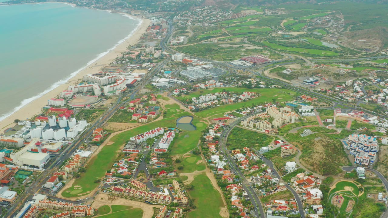Opening aerial frame over San Jose del Cabo showing pale ocean, sandy shore, green golf fairways, resort buildings, red-roofed homes, roads, hazy hills, and no visible people.