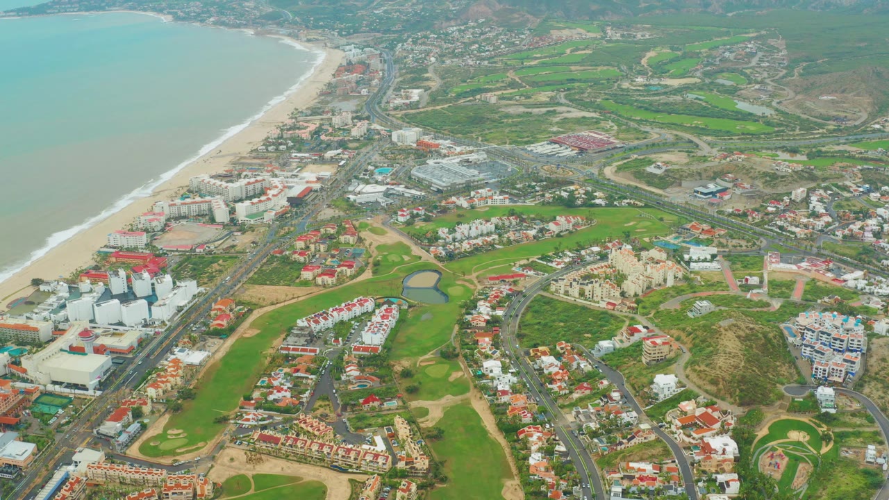 San Jose del Cabo aerial frame showing a long sandy shoreline, pale water, a green golf course with curving fairways, red-roofed homes, white apartment-like buildings, roads, and no visible people.