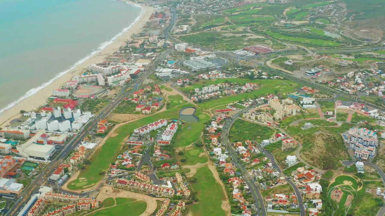 Aerial view in San Jose Del Cabo with the beach and surf running along the left side, green fairways in the center, and residential streets extending inland, with no visible people.