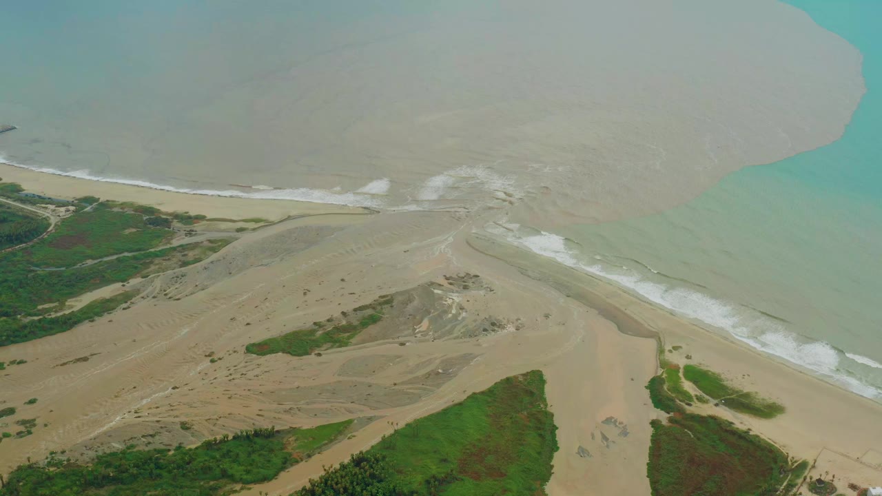 Aerial frame from San Jose del Cabo, Baja California Sur showing the sandy river mouth, pale brown water plume, white wave lines, and green coastal vegetation, with no visible people.