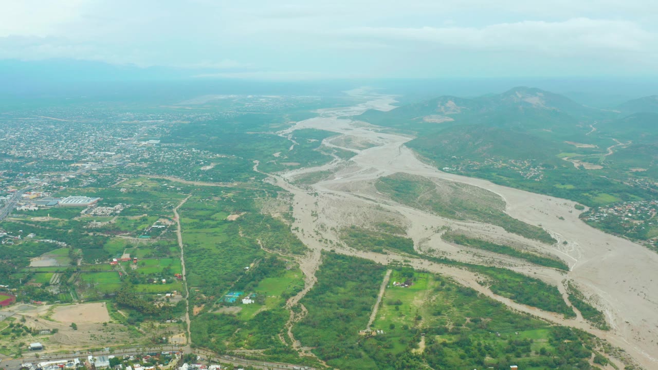 Opening aerial frame over San Jose del Cabo BCS Mexico showing a pale braided riverbed through green valley land, a town in the distance, mountains on the horizon, and no visible people.