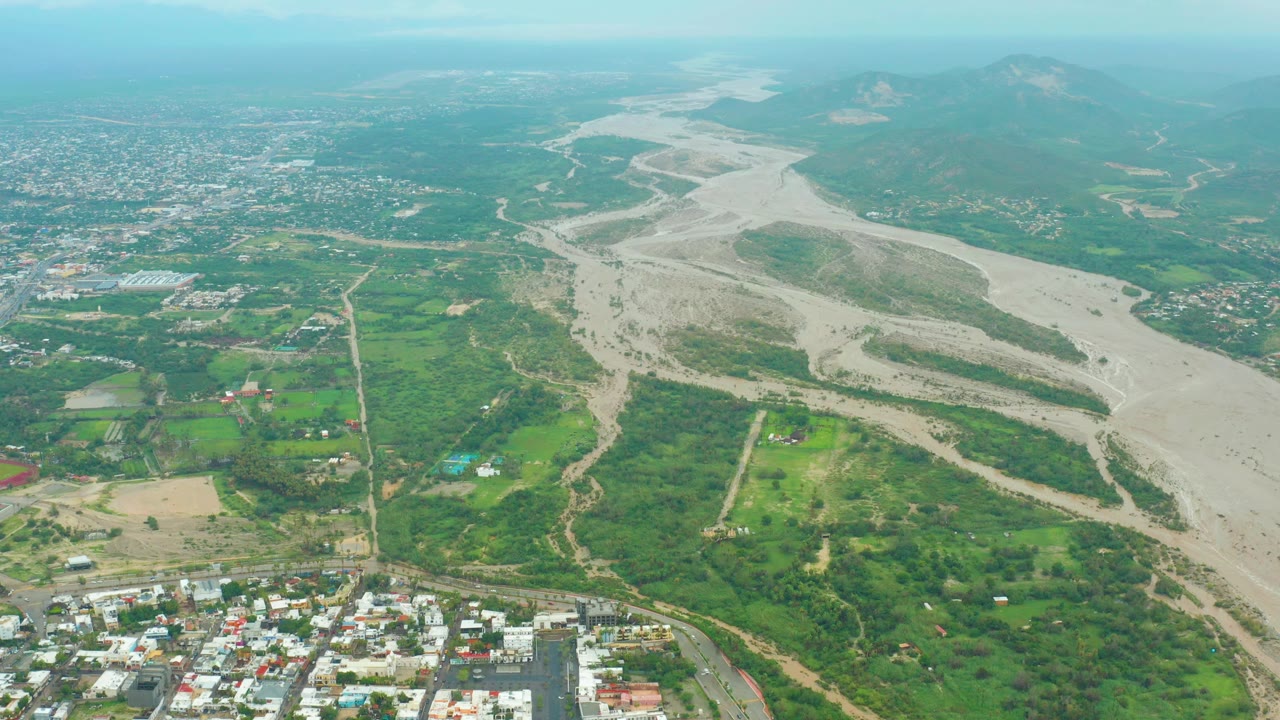 Opening aerial frame over San Jose del Cabo, Baja California Sur, showing a pale dry riverbed between green fields and urban development, with distant mountains and no visible people.