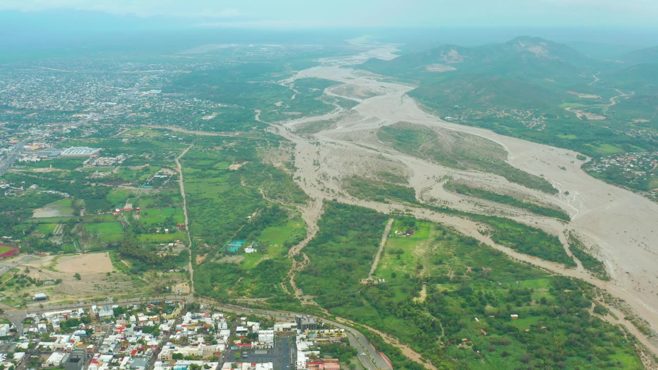San Jose del Cabo aerial view with a broad sandy riverbed cutting through green vegetation, a denser city area beyond it, mountain silhouettes, and no visible people.