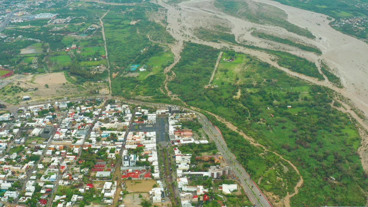Aerial frame from San Jose del Cabo, Baja California Sur, showing a pale muddy riverbed at upper right, green terrain in the middle, town blocks at lower left, and no visible people.