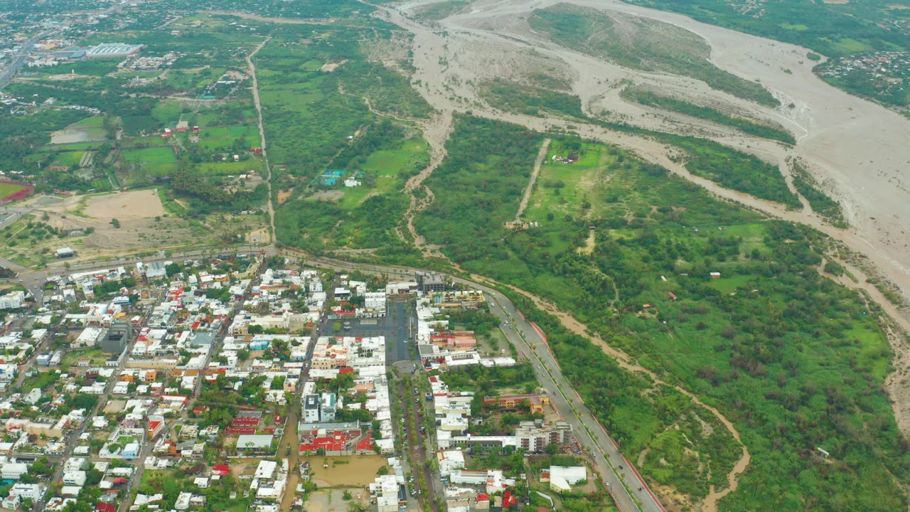 At 0:02, the aerial frame shows the braided muddy riverbed across the upper right, greener land through the center, and a compact urban grid at the bottom, with no visible people.