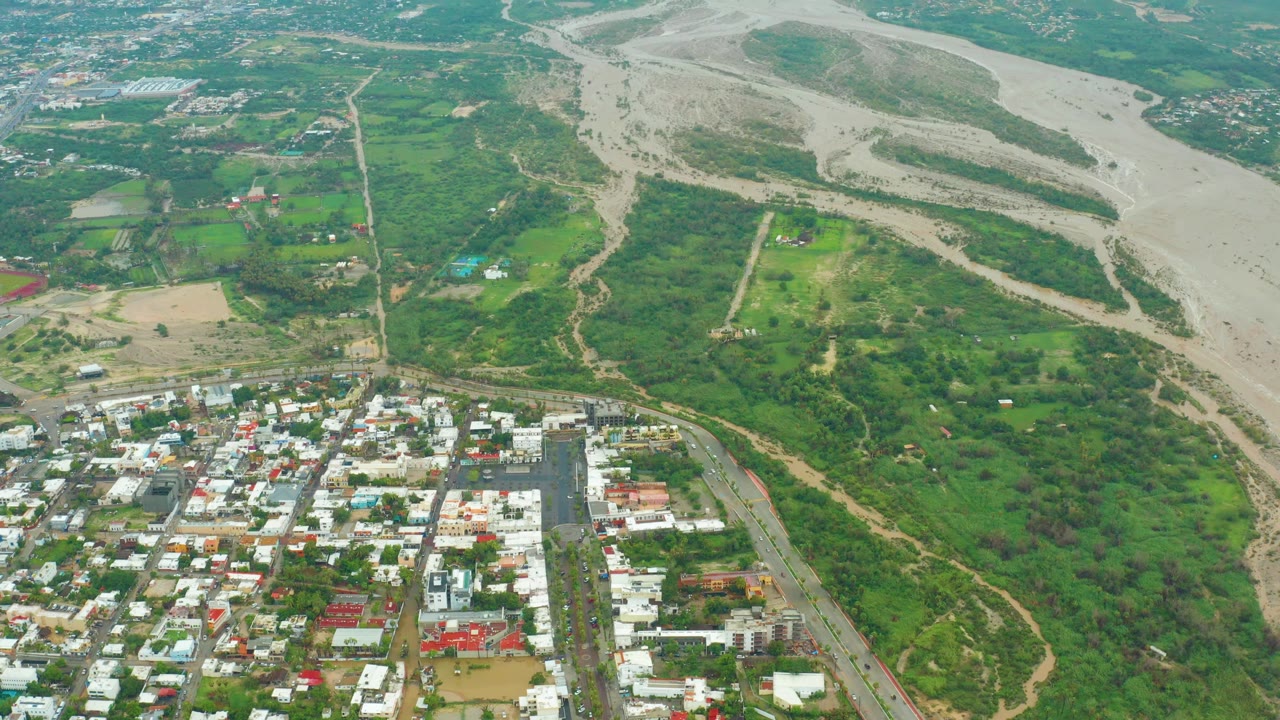 Drone-like aerial over a green wash, muddy riverbed, and low buildings.