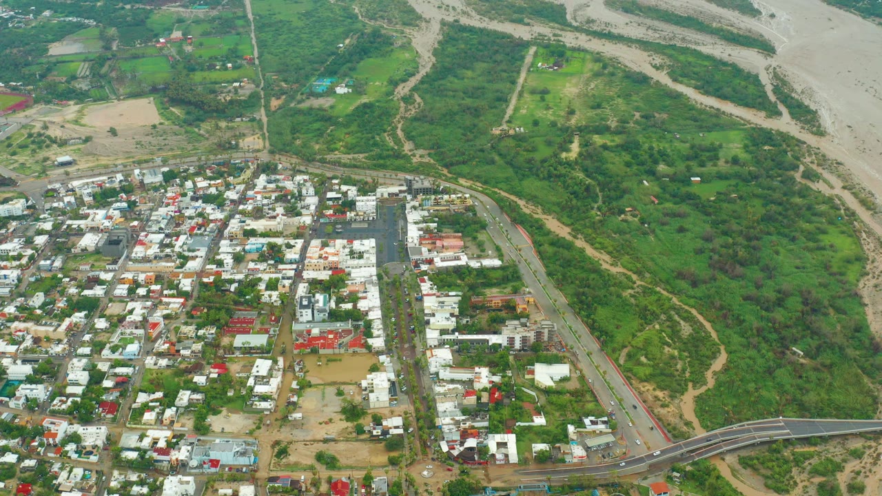 High aerial view in San Jose del Cabo, Baja California Sur, showing low town buildings, a main road, green riverbank vegetation, a broad exposed riverbed, and no visible people.