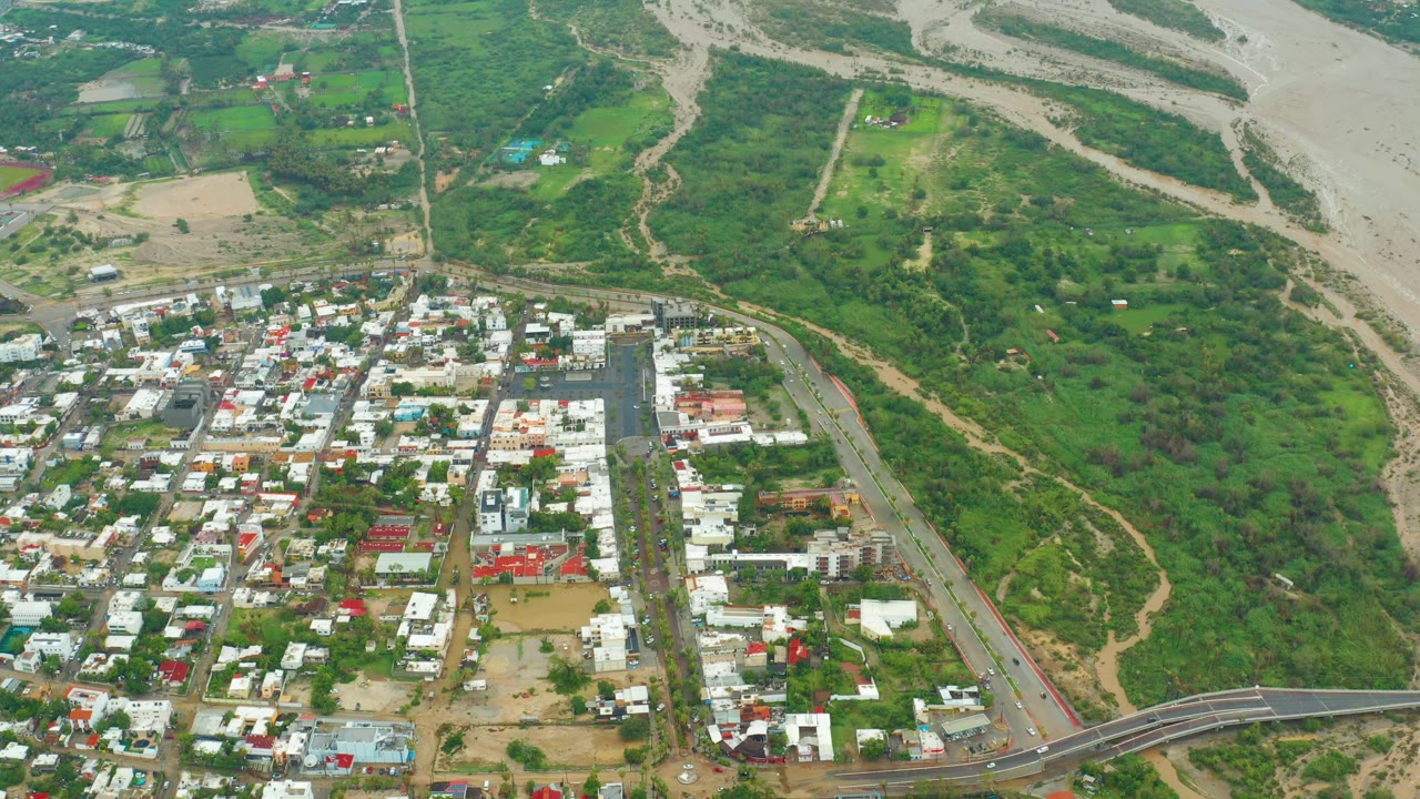 High oblique aerial view in Baja California Sur with town blocks in the lower half, dense green vegetation across the middle, a pale riverbed in the upper frame, and no visible people.