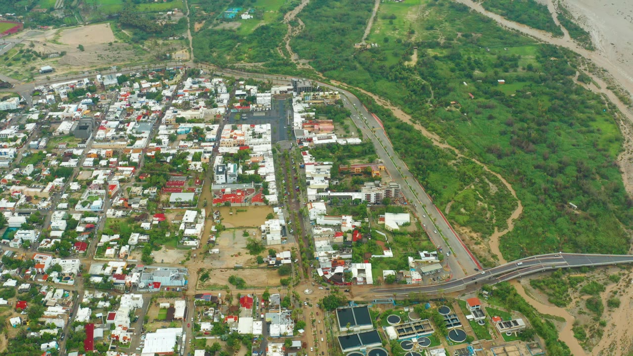 Aerial frame from San Jose del Cabo, Baja California Sur showing town blocks, a bridge, green hills, and a pale exposed riverbed, with no visible people.