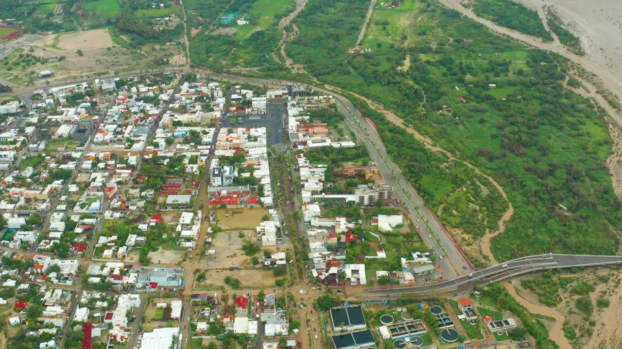 San Jose del Cabo aerial frame showing compact buildings and roads below, a bridge near the bottom, and green terrain leading into an exposed riverbank, with no visible people.