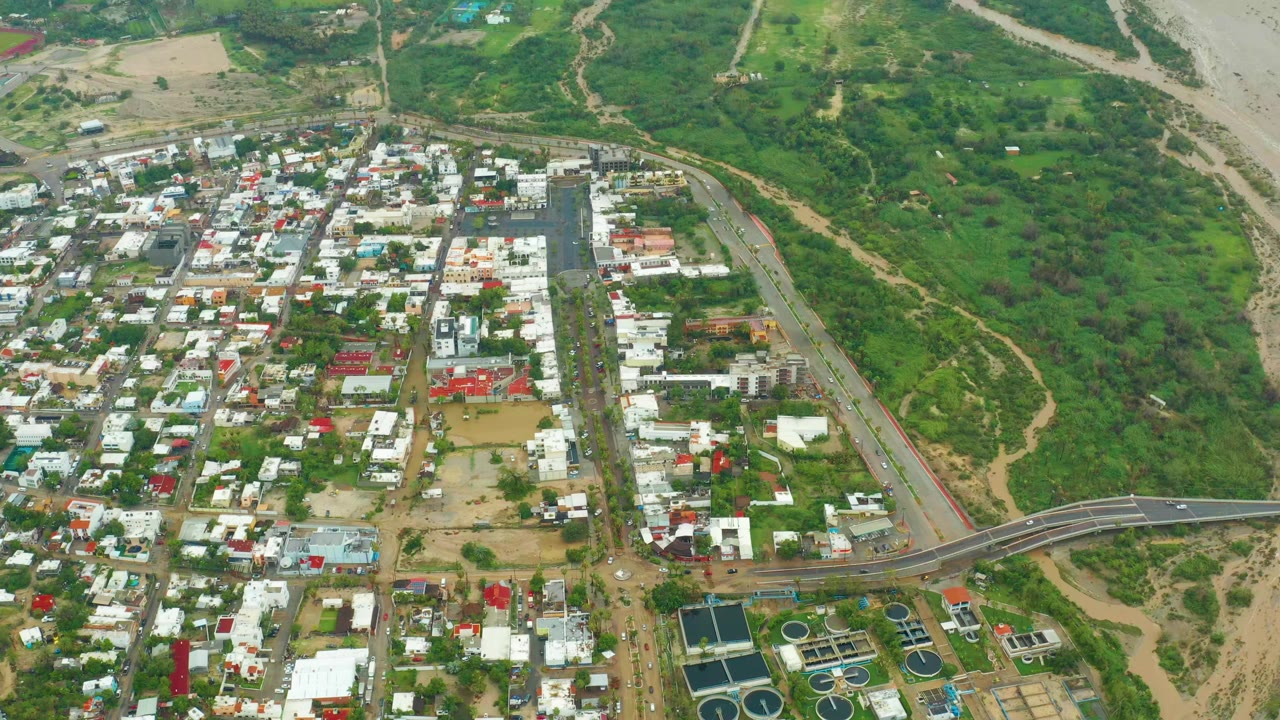 Opening aerial frame over San Jose del Cabo, Baja California Sur, with residential blocks on the left, a curving road and bridge below, green land to the right, and no visible people.
