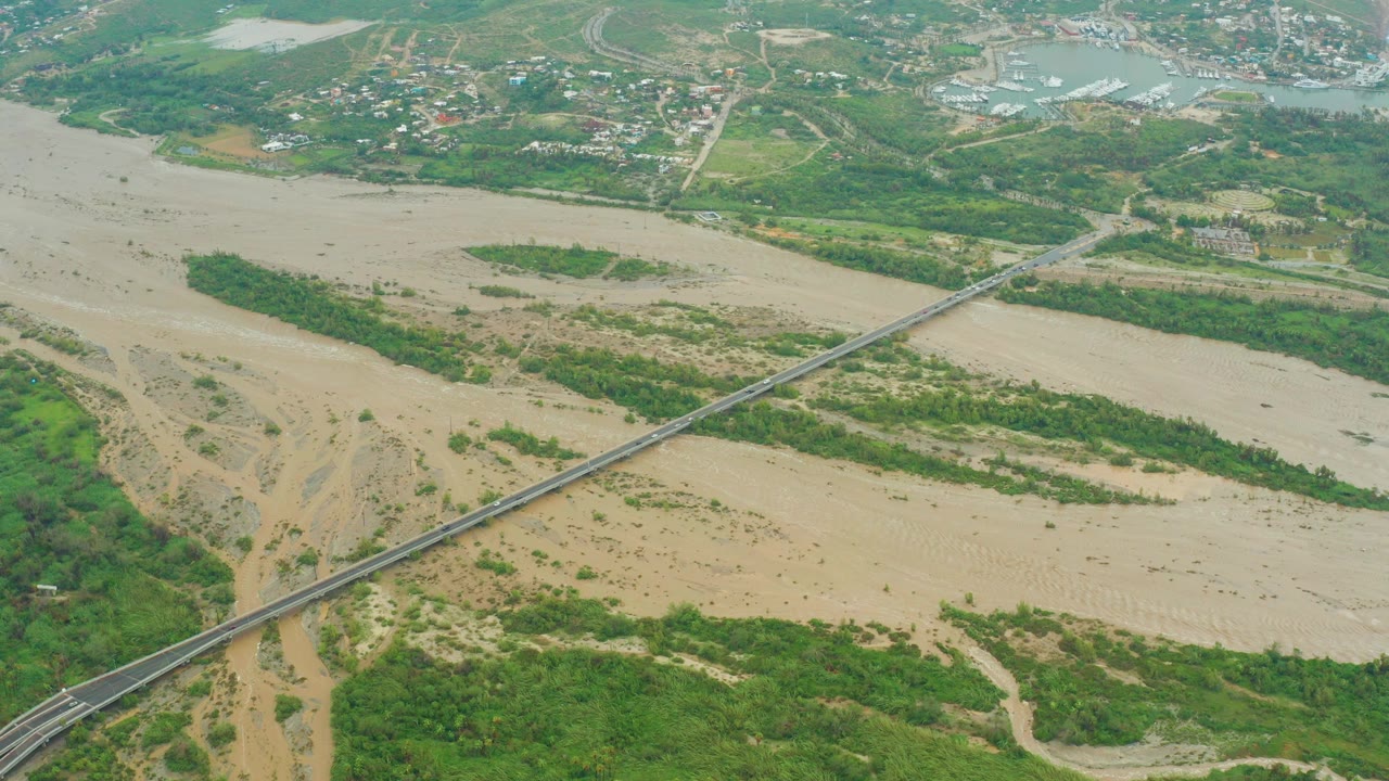Aerial view in Baja California Sur, Mexico, showing a muddy riverbed with exposed sand, green banks, a diagonal road bridge, distant buildings, and no visible people.