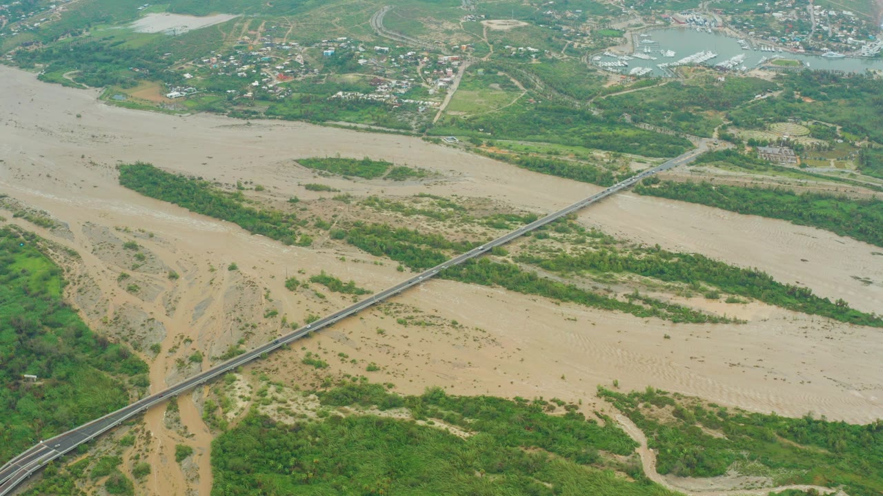 Aerial frame in Baja California Sur, Mexico, showing tan mud and sand patterns across the riverbed, a long bridge cutting diagonally through the scene, green banks, distant roads, and no visible people.