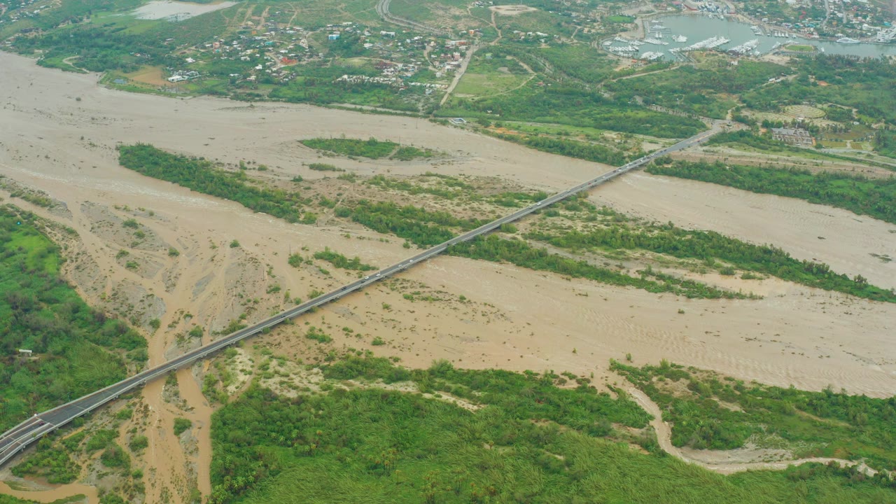 Aerial view in San Jose del Cabo, Baja California Sur, showing a muddy riverbed with green islands, a diagonal bridge, distant roads and buildings, and no visible people.