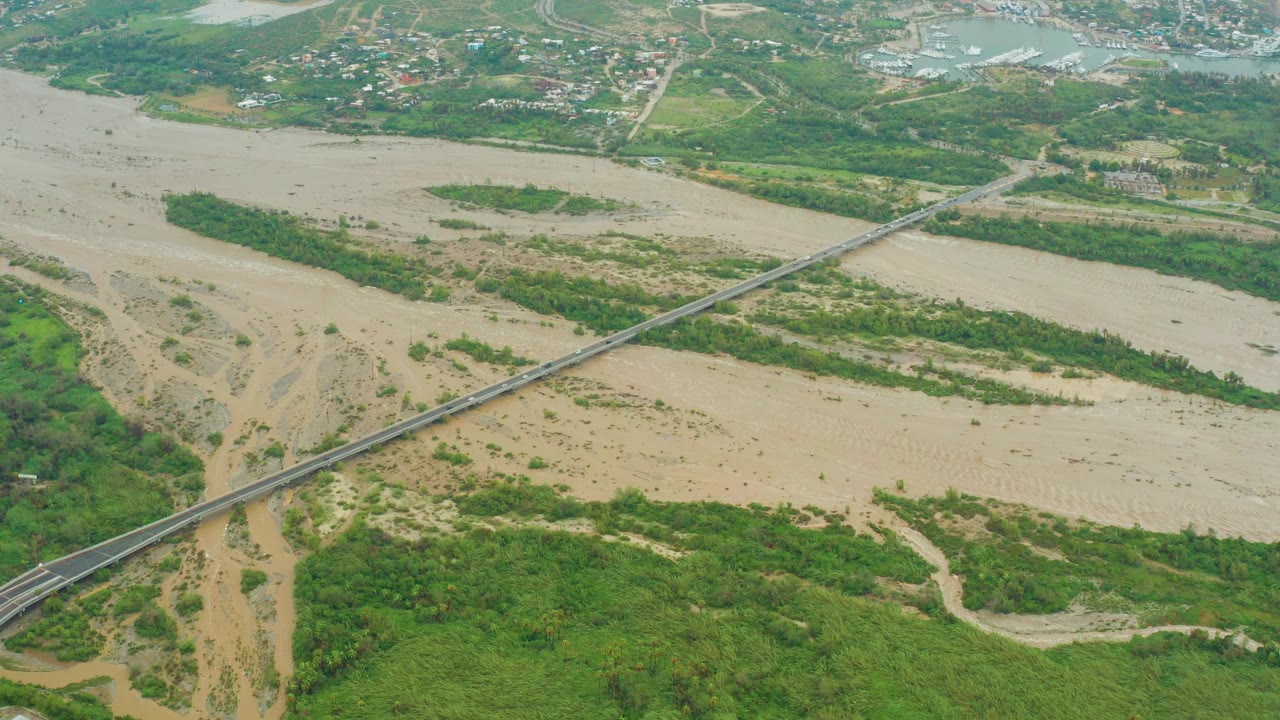 Aerial frame from Baja California Sur, Mexico, showing muddy water spread across a wide floodplain, a long road bridge, vegetated banks, pale sand deposits, distant development, and no visible people.