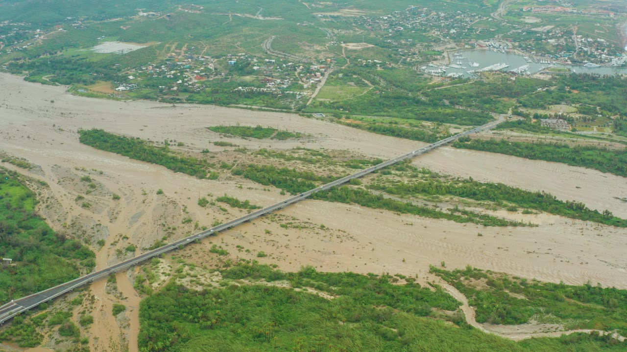 Aerial frame from San Jose del Cabo, Baja California Sur, showing a tan muddy river, diagonal bridge, green vegetation, distant roads and buildings, and no visible people.