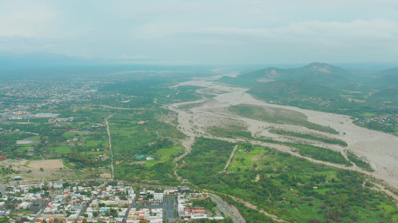 Aerial view in San Jose del Cabo, Baja California Sur, showing a pale riverbed, green fields, town buildings along the lower edge, hazy hills, overcast sky, and no visible people.