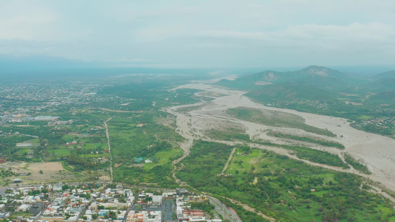 Drone-style aerial view in San Jose del Cabo showing green valley land around a pale winding riverbed, town blocks near the bottom edge, mountains beyond, overcast light, and no visible people.
