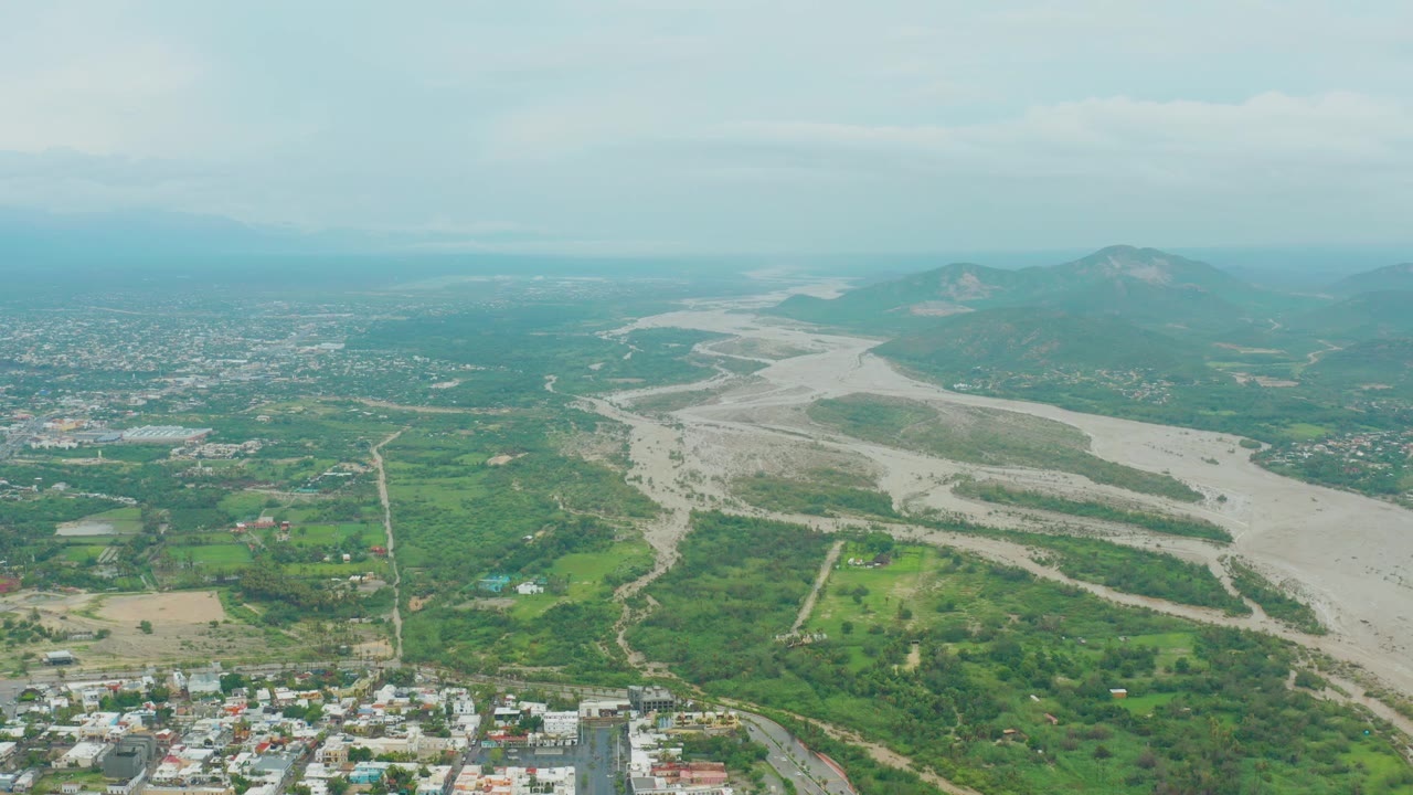 Aerial view across a broad riverbed, fields, scattered buildings, and hazy hills.