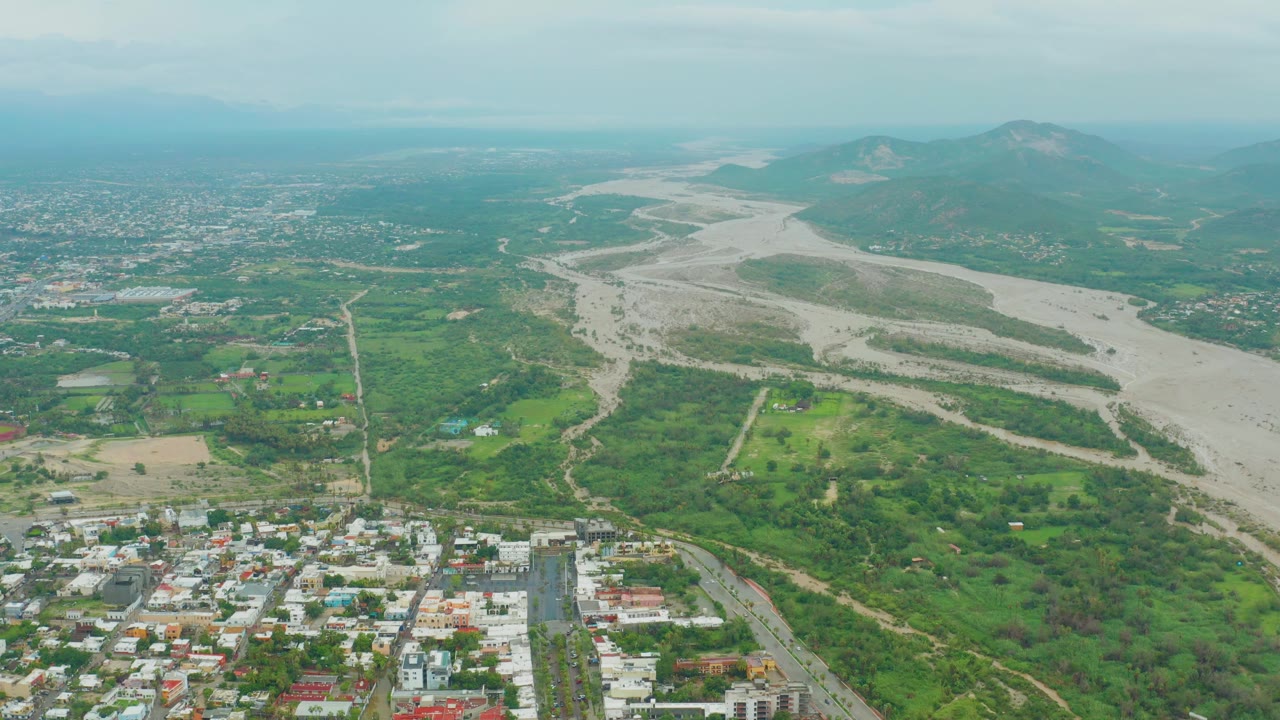 A broad aerial frame of San Jose del Cabo shows the river channel crossing the valley, with dense green vegetation, small buildings in the foreground, and mountains under cloud cover, no visible people.