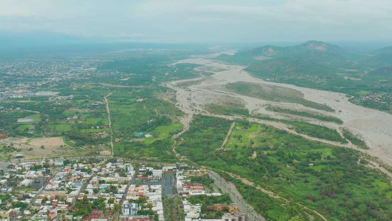 San Jose del Cabo appears from above with town blocks at the bottom, a light-colored riverbed sweeping through green valley land, and distant mountains softened by haze, no visible people.