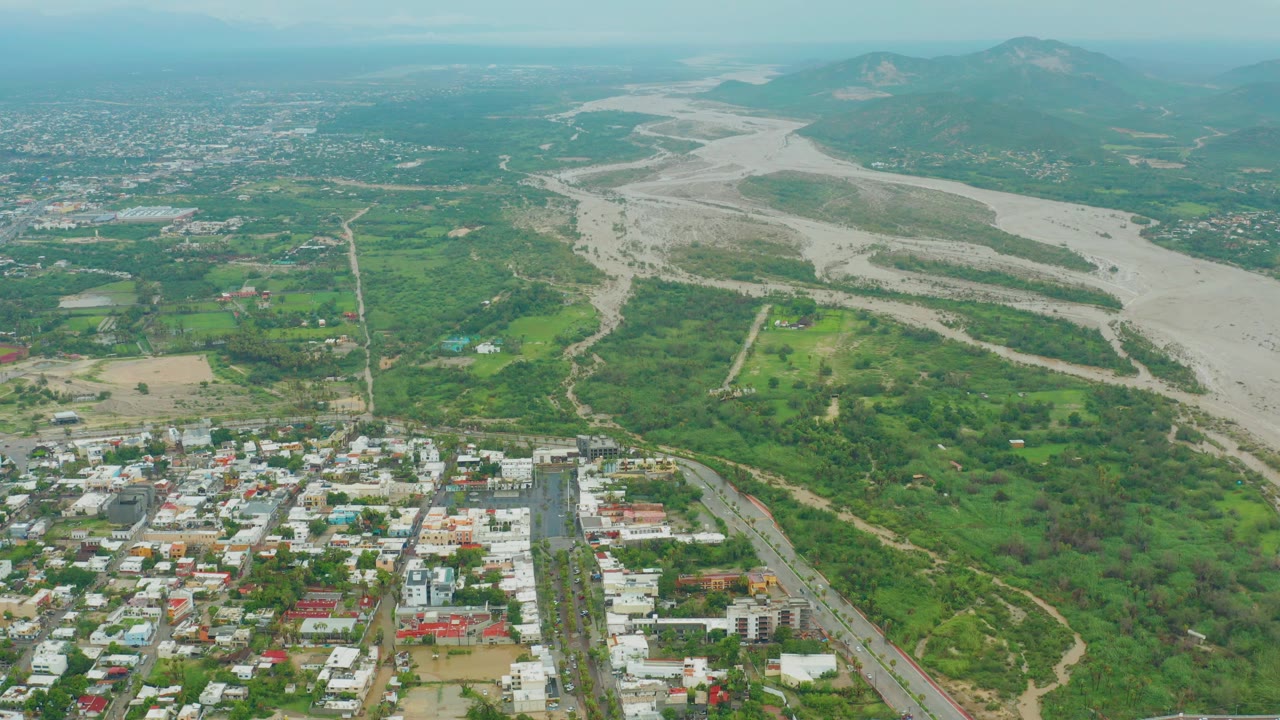 Opening aerial frame over San Jose del Cabo BCS Mexico with town rooftops in the foreground, a broad pale riverbed through green land, distant mountains, and no visible people.