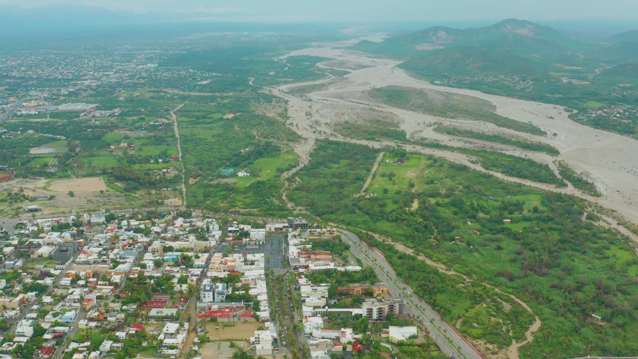 Drone view of a dry river channel running past green land and urban buildings.