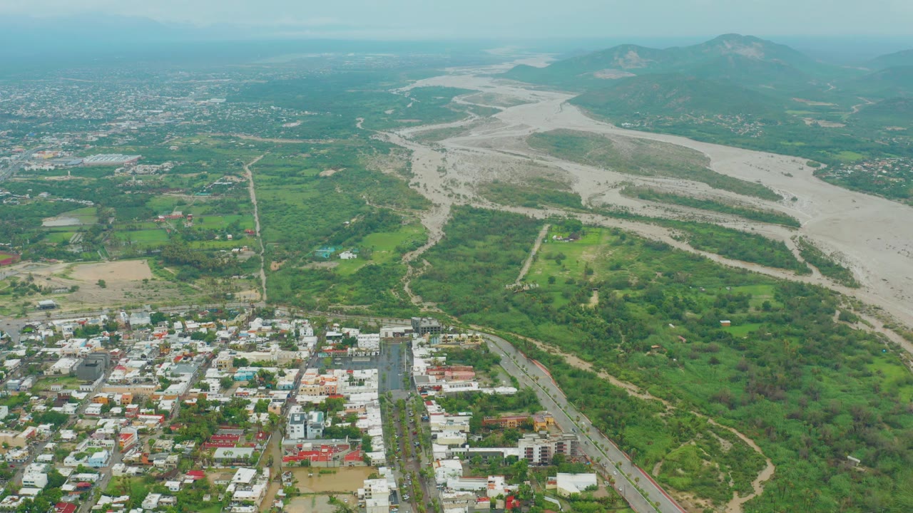 San Jose del Cabo BCS Mexico aerial with foreground neighborhood roofs, rectangular field patches, a pale river course across the middle distance, mountains beyond, and no visible people.