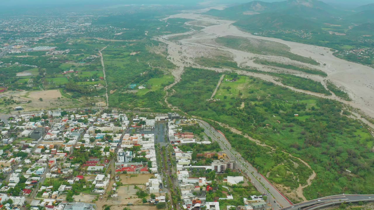 Opening aerial frame over San Jose del Cabo, Baja California Sur, with low-rise town blocks in the foreground, a broad pale riverbed beyond green terrain, distant hills, and no visible people.