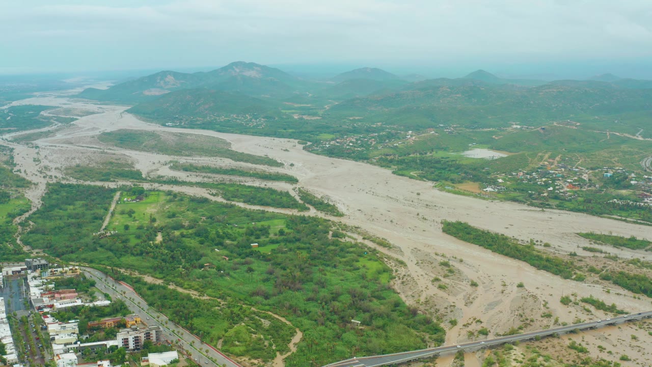 Wide aerial over a pale muddy river channel bordered by green vegetation and low urban development.