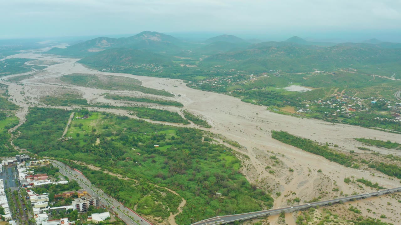 Aerial frame with the muddy riverbed, green patches, nearby buildings, and a road or bridge at the bottom.